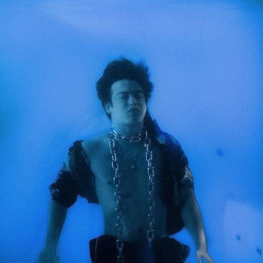 A young man submerged in blue water, wearing a chain necklace and a black shirt, creating an artistic underwater scene.